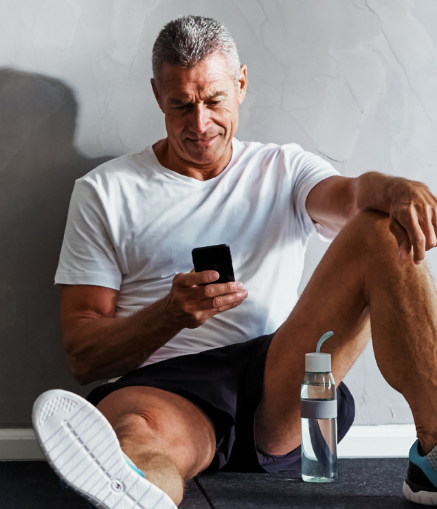 Middle-aged man resting after a workout, sitting on the floor and checking his smartphone, with water bottle nearby, representing fitness recovery and health tracking.